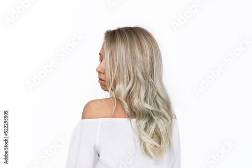 A young charming blonde woman with wavy blond hair in a white blouse standing with her back isolated on a white background. Result of coloring, highlighting, perming. Beauty and fashion