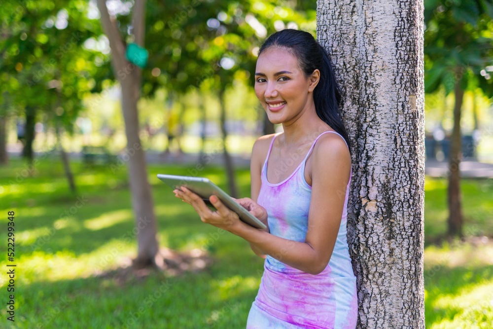Portrait photo of the beautiful moment of a young asian beautiful lady with colorful dress happily and relaxingly using her digital tablet under a tree at a garden park Portrait photo of the beautiful moment of a young asian beautiful lady with colorful dress happily and relaxingly using her digital tablet under a tree at a garden park