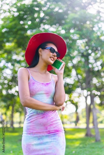 Portrait photo of the beautiful moment of a young asian beautiful lady with red hat and sunglasses happily chatting on her phone with her friends during a garden park strolling