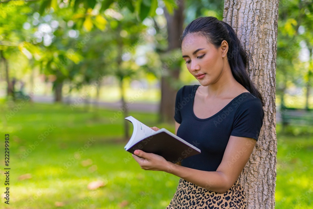 Obraz premium Portrait photo of the moment of a young beautiful asian female lady reading a book under a tree in an outdoor park during her summer holidays