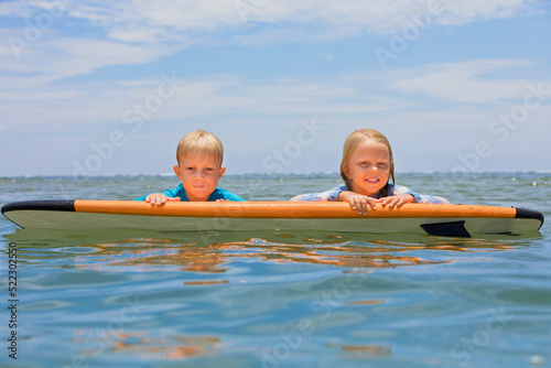 Happy baby boy and girl - young surfers ride with fun on one surfboard. Active family lifestyle, kids outdoor water sport lessons, swimming activity in surf camp. Sea beach summer holiday with child. © Tropical studio