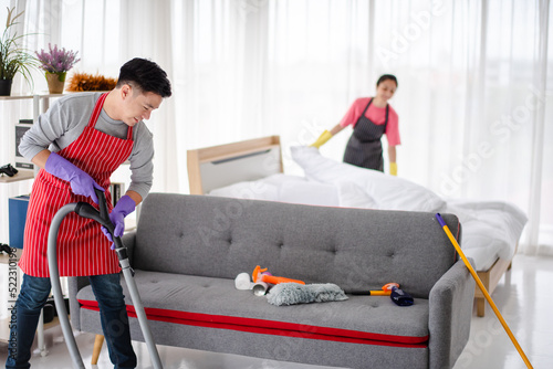 Young Asian family, Husband and wife are cleaning the house together. he is vacuuming,  using a vacuum cleaner, and the wife is wearing the same apron but is black. She is changing new blankets