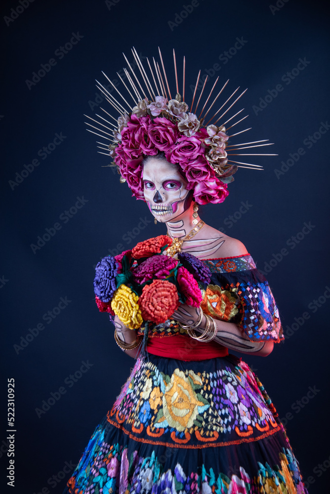 catrina woman dressed in a mexican chiapas costume with a black background pink rebozo and skull and bones makeup on her hands