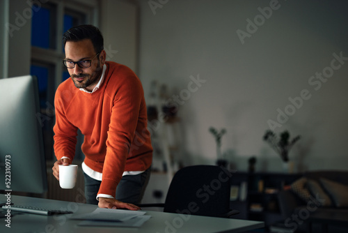 A man hardworking at the office, staying overtime, drinking coffee.