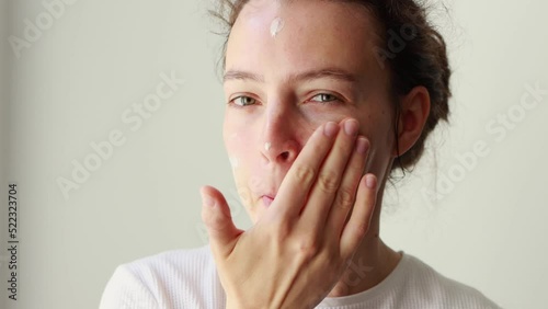 Happy attractive young caucasian woman applying skincare cream on face, looking at camera, putting soft hydrated moisturizing lifting facial creme. Beauty and healthy skin care treatment concept.