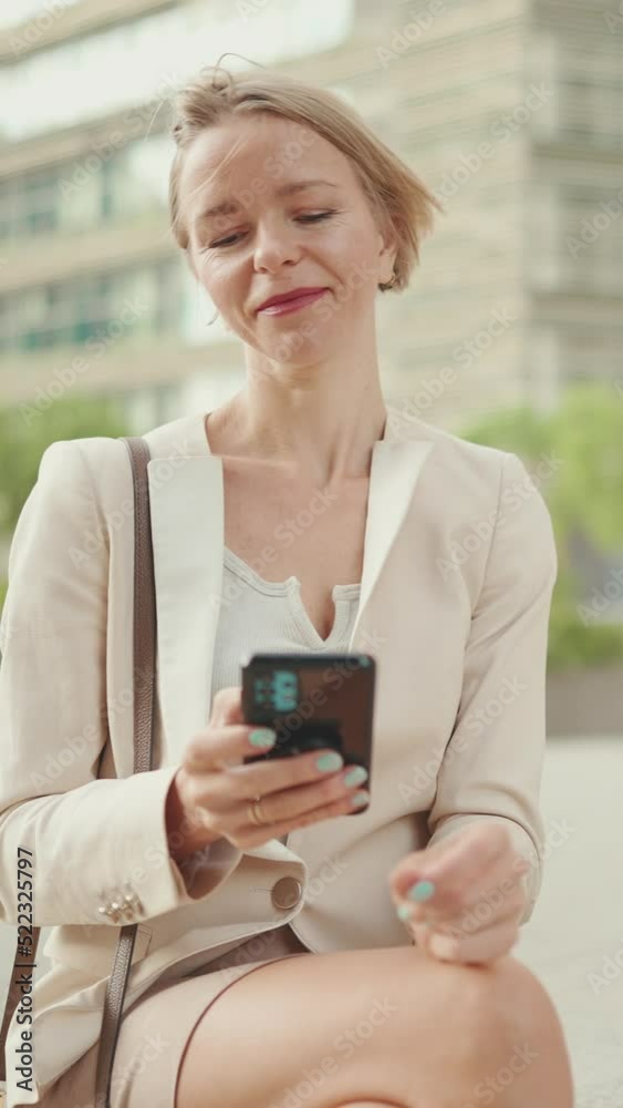 VERTICAL VIDEO: Smiling woman with blond hair wearing beige suit using mobile cellphone sitting outside on the street