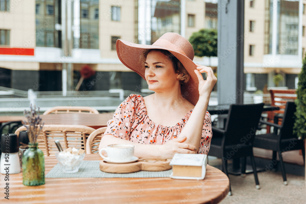 Outdoor fashion portrait of a stunning woman sitting in a cafe. I drink coffee and read an old book. a woman in a dress and a hat.