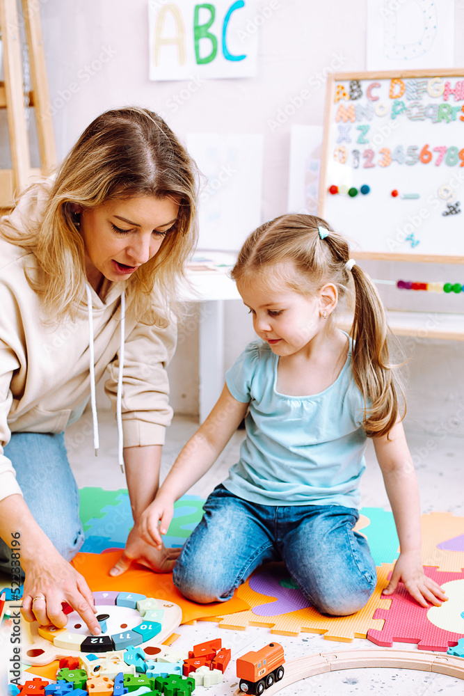 Fototapeta premium Little girl wear T-shirt, jeans sitting with young woman teacher teaching how to play with parts of clock with numbers.