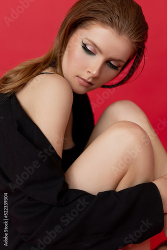 Portrait of young beauteous woman with ginger hair wearing black swimsuit, cardigan with bare shoulder, hugging knees.