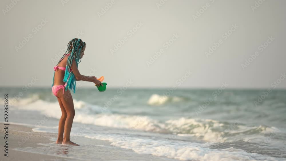 A girl with african braids in a summer costume plays on the beach with shells near the sea with waves under sunny sunset