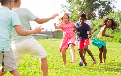 Happy preteen girls and boys of different nationalities playing football on green lawn in spring city park.. © JackF