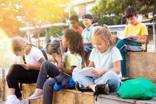 Multiethnic group of schoolchildren sitting with workbooks in schoolyard in warm autumn day.