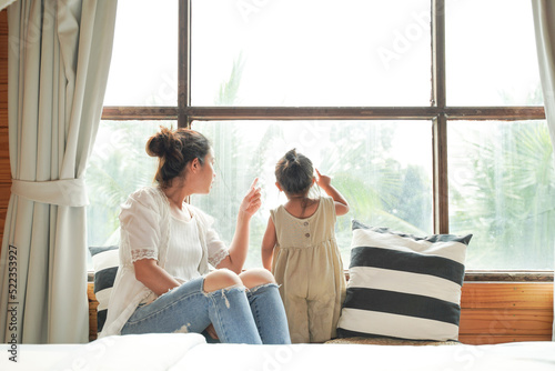 Happy couple of mom and child girl. The family looks through the house windows on the garden, the mother shows a finger in the window and the daughter watches.