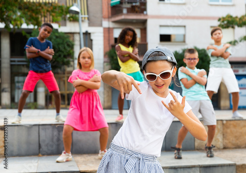 Modern preteen girl krump dancer wearing stylish sunglasses and cap posing during performance with group on summer city street