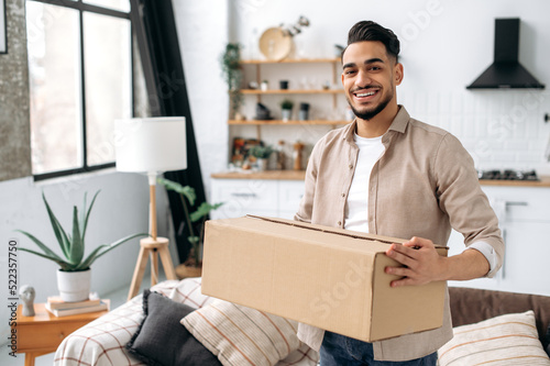 Happy confident indian or arabian guy, stand at home in living room, holding a large cardboard box, received a long-awaited parcel from the online store, preparing to unpack, looks at camera, smiles © Kateryna
