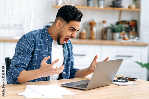 Angry annoyed young man, Arabian or Indian company worker, freelancer, stylishly dressed, working from home, sitting in the kitchen at a desk, yelling at the laptop, dissatisfied with the result