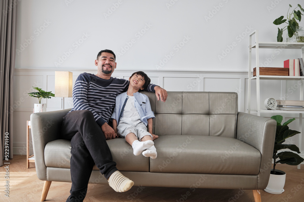 Portrait of Asian Thai family, one adult dad and little son happiness home living together, relaxing and smile, leisure on sofa in house's white room, lovely weekend and wellbeing domestic lifestyle.