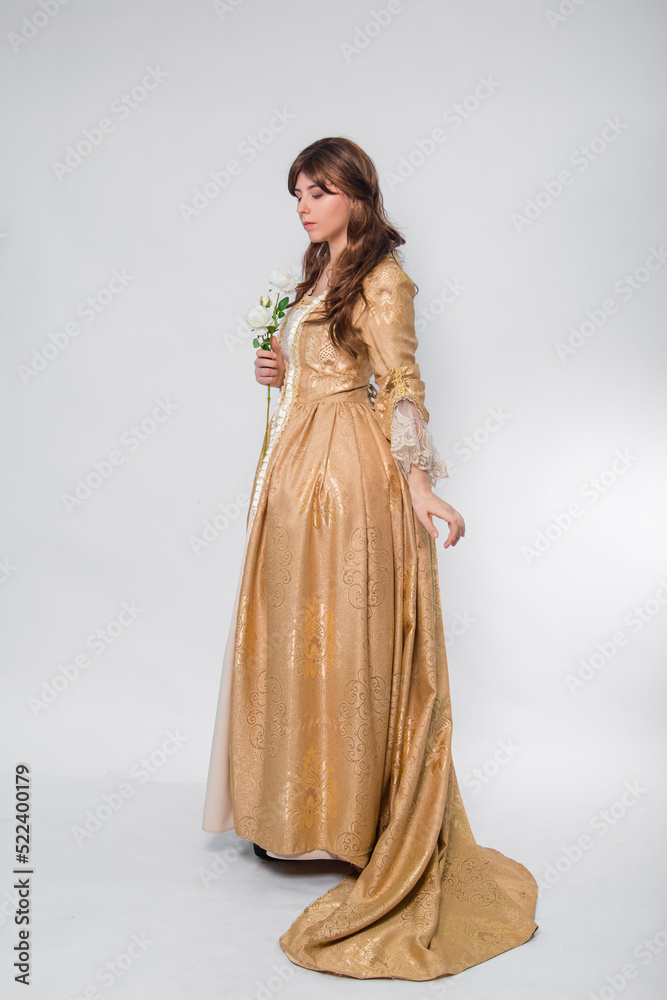 Fototapeta premium Full length portrait of a woman in a gold dress in the style of the rococo era, standing and posing with a flower in hand isolated on a white background.