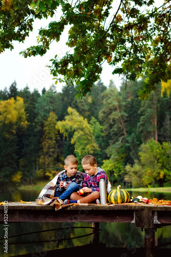 The boys are sitting with a thermos by the river on a wooden pier. Friends on a picnic in nature in autumn. The children wrapped themselves in a blanket. © Elena 