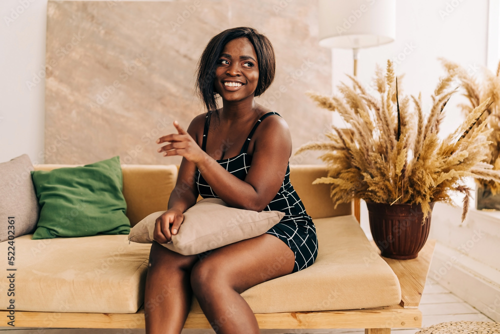 Naklejka premium Happy stylish beautiful female student with hair looks at camera stands in cozy home interior. Smiling attractive young woman wears black dress headshot portrait.