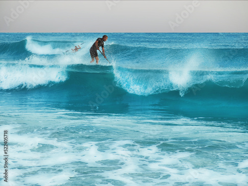 Men are SUP-surfing during a storm, one is standing on the board, the other is sitting. Steep waves, cloudy skies, athletes catching waves. Splashes of water. Sea waves, landscape, water sports