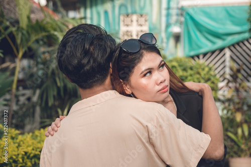 An unsure asian woman hugs her partner faking forgiveness or showing doubt. An emotionless face concealing her inner conflict. Outdoor scene. photo