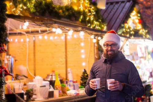 Close-up of a man in a Merry Christmas fair in the city hot drinks  enjoying  dressed warm. The man is wearing a winter jacket  a Santa Claus hat  and eyeglasses. Cocept  Christmas Winter Vacation