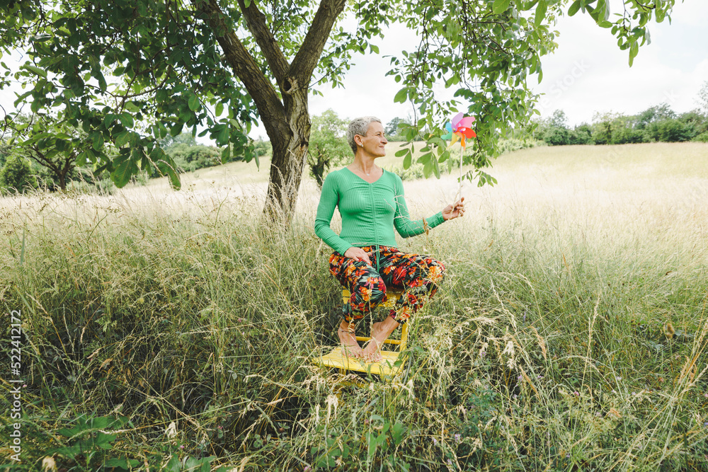 Obraz premium Middle aged woman with short light hair, green top and colorful flower pants in high meadow holding colorful pinwheel