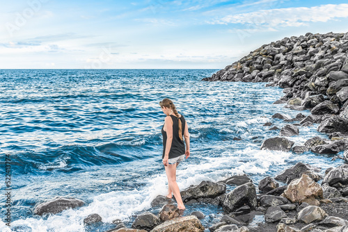 A young woman enters the ocean from a stone shore - view from the back. A girl with a pigtail in white shorts and a black T-shirt stands in sea water against the backdrop of waves © ioanna_alexa