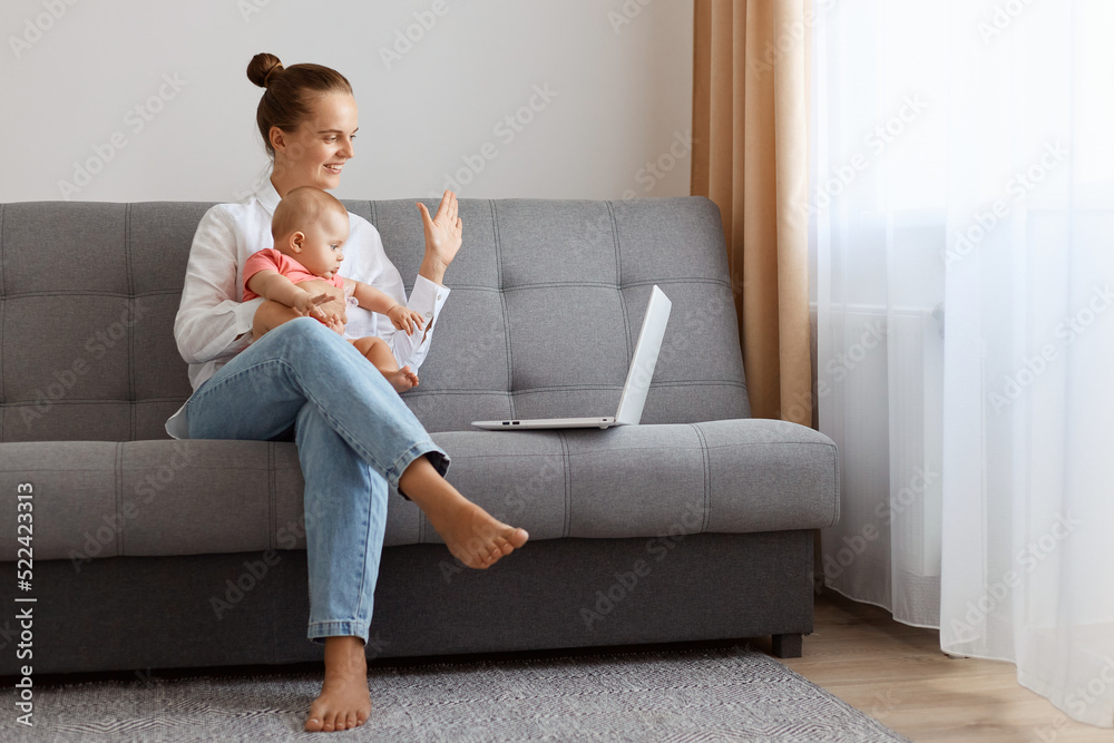 Indoor shot of smiling woman in white t shirt sitting on sofa with baby daughter and working on laptop, having video call, saying hi and showing hello gesture with hand. Indoor shot of smiling woman in white t shirt sitting on sofa with baby daughter and working on laptop, having video call, saying hi and showing hello gesture with hand.