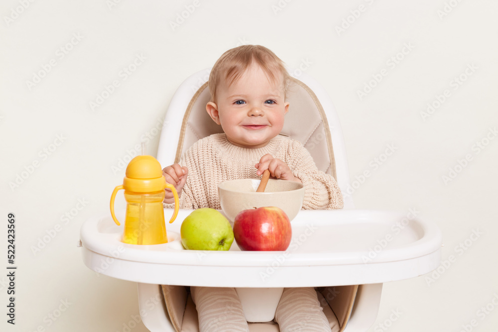 Portrait of smiling baby wearing beige sweater sitting in a child's chair, isolated on a white background, holding spoon and eating vegetable or fruit puree. Portrait of smiling baby wearing beige sweater sitting in a child's chair, isolated on a white background, holding spoon and eating vegetable or fruit puree.