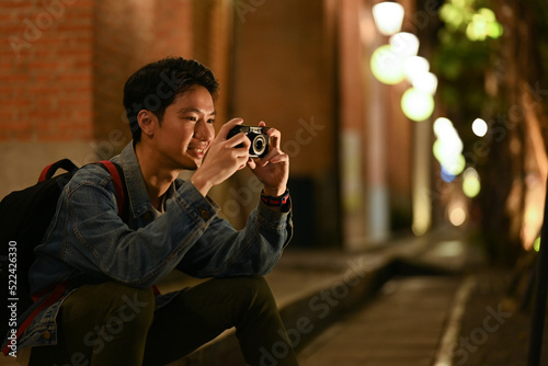 Young male traveler taking photo with his camera, sitting on stairs in the night city with blurred night street lights background