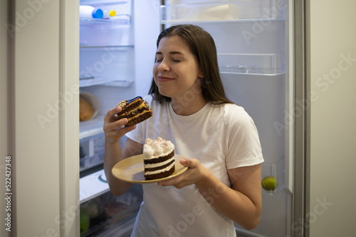 Woman smiling enjoys a delicious sweet chocolate cake at night alone near the fridge. Funny photo. A woman with trouble eating. Carbohydrates  calories. Nocturnal Harmful Forbidden Food