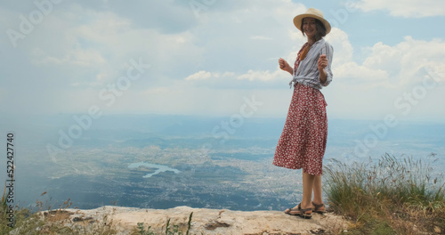 Happy traveler on mountain top. Carefree woman traveler in hat, red dress dancing, jumping on mountain, backdrop of Tirana city, Albania. Happy person enjoying travel by country. Concept of wanderlust