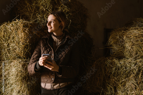 Fototapeta Young jockey woman using mobile phone while standing by hay in stable