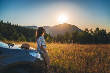 Woman traveler looking at the sunset in the field in the mountains standing near car