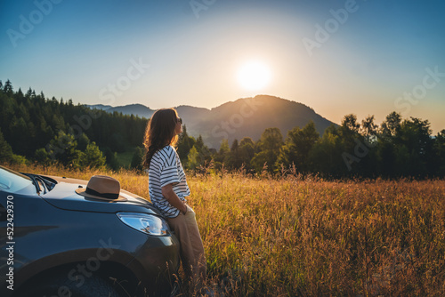 Woman traveler looking at the sunset in the field in the mountains standing near car
