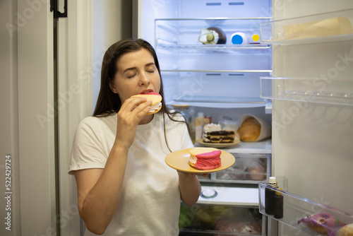 Woman in the sleepwear eating sweet donuts near the refrigerator. Concept: breaking the diet night eating high calorie. Stop diet and gain extra pounds due to carbs food and unhealthy eating