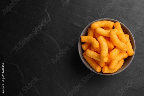 Many tasty cheesy corn puffs in bowl on black table, top view. Space for text