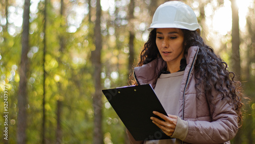 Foto Millennial female technician ecologist looking up at treetops, Young indian woman in hardhat with clipboard taking measures checking trees