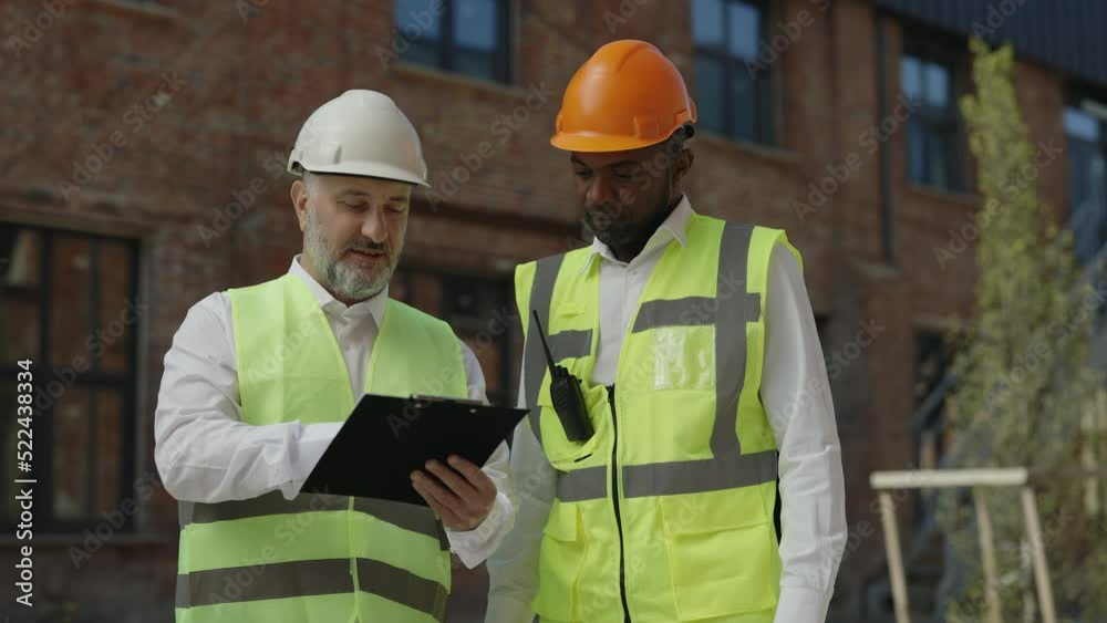 Diverse factory workers shaking hands after discussion of progress in building construction outdoors. Foreman and engineer in safety uniform holding clipboard with approved plan of actions. Diverse factory workers shaking hands after discussion of progress in building construction outdoors. Foreman and engineer in safety uniform holding clipboard with approved plan of actions.