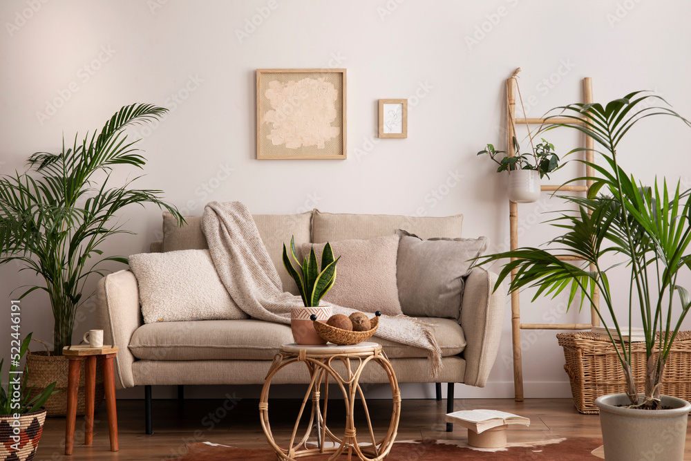 Fototapeta premium Boho and cozy interior of living room with design beige sofa, pillows, mock up poster frames, rattan coffee tables, plants, bamboo ladder, decoration and personal accessories. Home decor. Template.
