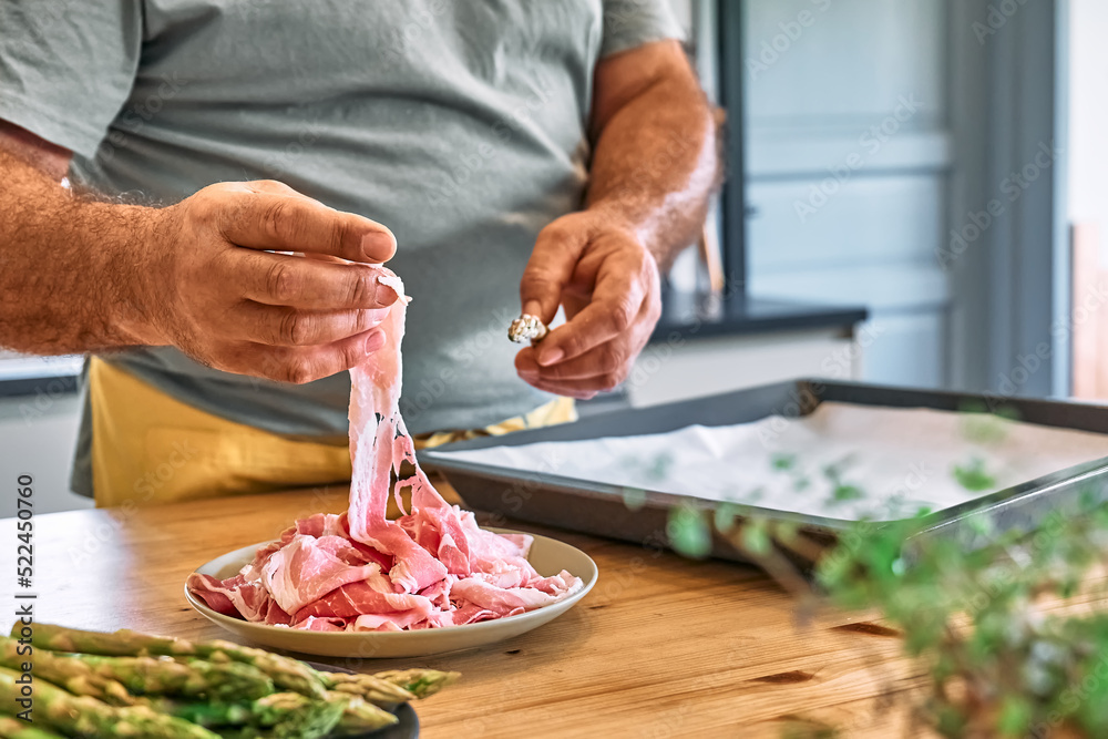 Man preparing young green asparagus sprouts wrapped in bacon on wooden table in the kitchen. Man's hand wrapping asparagus in ham for baking.