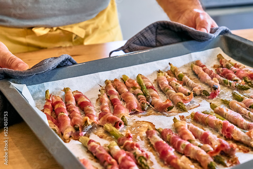 Man blogger preparing young green asparagus sprouts wrapped in bacon on wooden table in the kitchen. Hands of man holding baking tray with asparagus wrapped in ham baked in oven.