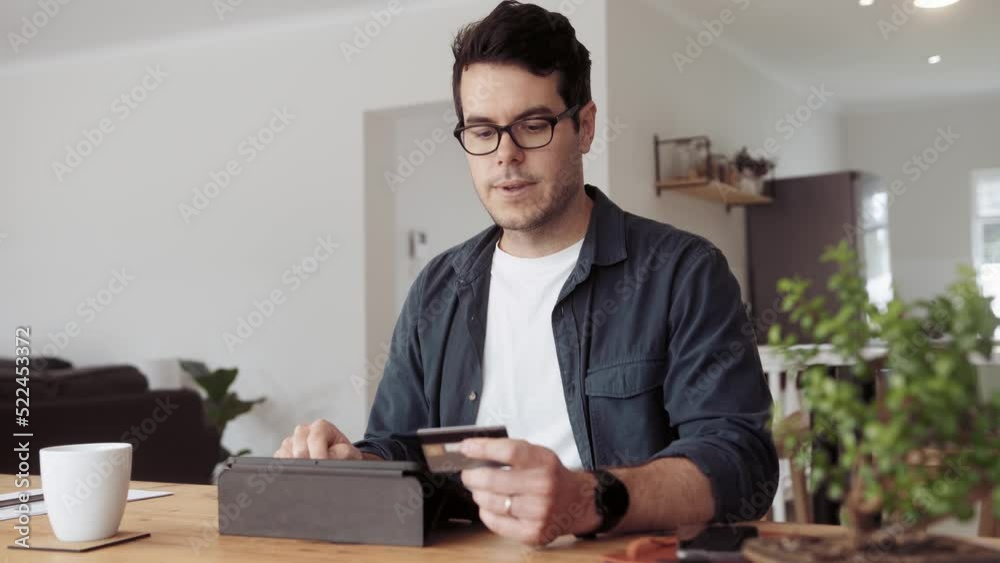 Caucasian man typing his card details onto an app on his tablet while sitting at his dining room table at home. Caucasian man typing his card details onto an app on his tablet while sitting at his dining room table at home.