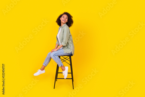 Full length profile photo of brunette lady sit on chair look up wear shirt jeans boots isolated on yellow background