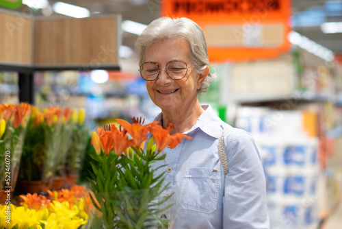 Senior caucasian woman at the supermarket selecting an orange flower plant in special offer, attentive at the price increase and inflation