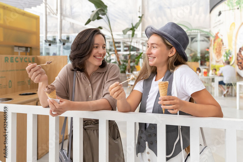 Happy female friends eating ice cream at food court and talking. Smiling teenage friends meeting in shopping mall. Female friendship concept