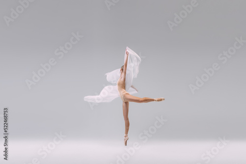 Portrait of young ballerina dancing with transparent fabric isolated over grey studio background. Standing on tiptoe on pointe