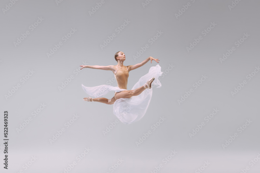 Portrait of young ballerina dancing with light transparent fabric isolated over grey studio background. Flying like a bird. Freedom